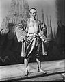 Black and white photo of a man with a shaven head in silky Asian garb; his chest is exposed and his feet are bare; he stands with hands on hips, glowering at the camera