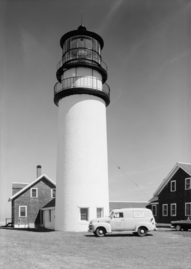 High-resolution black and white photo of the lighthouse and keeper's house. Parked in front of the lighthouse is a 1950s van, with a military emblem and the words "United States Coast Guard" stenciled on the driver's door, and on the side of the van is a military advertisement that reads "The U.S. Coast Guard NEEDS MEN! Join Now! No waiting".
