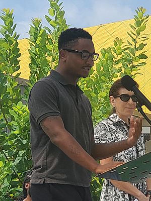 Ossé in a polo shirt and glasses speaking into a microphone outdoors on a sunny day