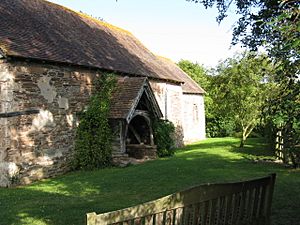 Part of the south face of a small stone church, seen from an angle, with tiled roofs and a gabled, wooden porch