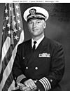 Top half of man in circa 1970 U.S. Navy officer uniform, before an American flag.