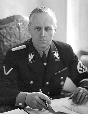 Portrait of a middle-aged man with short grey hair and a stern expression. He wears a dark military uniform, with a swastika on one arm. He is seated with his hands on a table with several papers on it, holding a pen.
