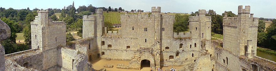 The courtyard of Bodiam Castle with the ruins of domestic buildings. In the centre of the image is the three-storey gatehouse. The curtain wall and the towers spaced along it are in good condition, contrasting with the interior. In the background, the landscape rises above the castle.