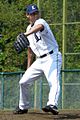 A man wearing a white baseball uniform with a navy blue "L" on the chest, a navy blue cap with a white "L" on the center, and a black glove on his left hand stands on the pitcher's mound in the midst of pitching a ball.