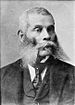 Head and shoulders of an older black man with short hair and extremely long, bushy sideburns connecting to a mustache. He is wearing a suit coat, vest, and tie.