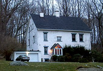 A two-and-a-half-story white house with blue trim and two white chimneys piercing the roof. A small garage is at its left with a car parked in front of it. There are bare trees in a wooded area behind it.