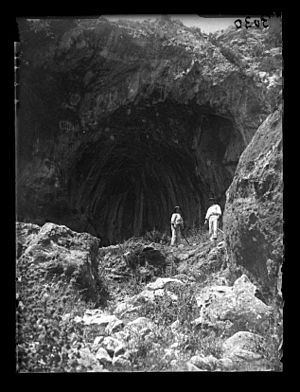 Black and white photograph showing the entrance to a cave, with two people standing in front, looking at the camera