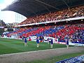 Photo of 7 cheerleaders performing on a football pitch in front of a packed stand with many on the lower tier waving red and blue flags.