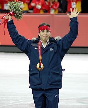 A man wearing a gold medal smiles with his arms raised above his head holding a flower bouquet in his left hand while wearing a dark blue tracksuit and a red bandanna on his head. There is a portion of the ice-rink in the background.